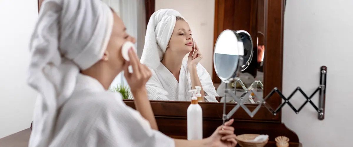 girl with a towel on her head and in a bathrobe posing smiling, sitting at a brown dressing table, looking in a retractable mirror mounted on the wall