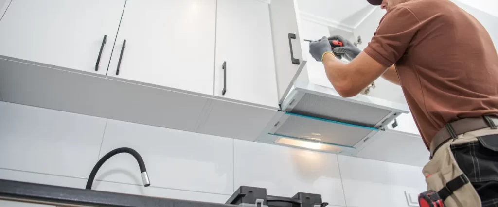 A professional contractor installing sleek white kitchen cabinets, using a screwdriver to secure hardware above a modern cooktop with a stainless steel range hood.